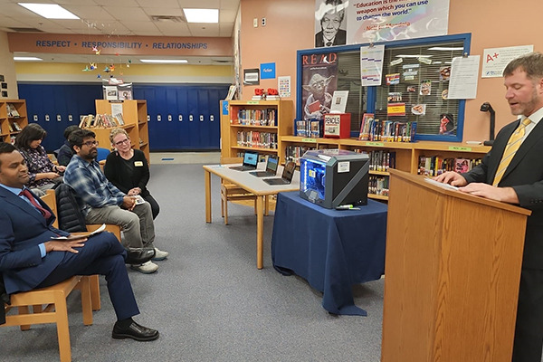 A speaker addressing attendees in a school library beside a 3D printer