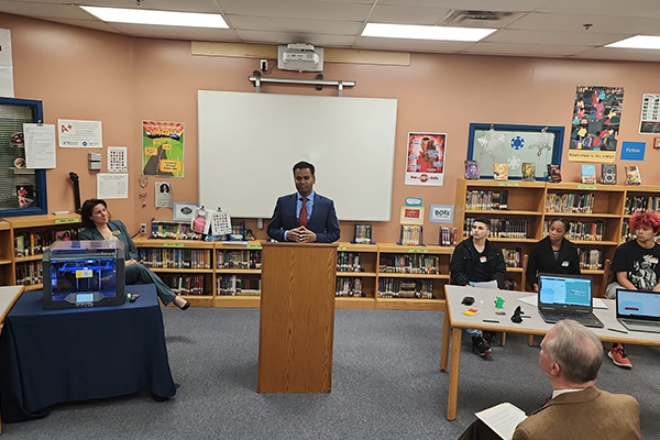 A speaker at a podium in a school library, with a 3D printer nearby